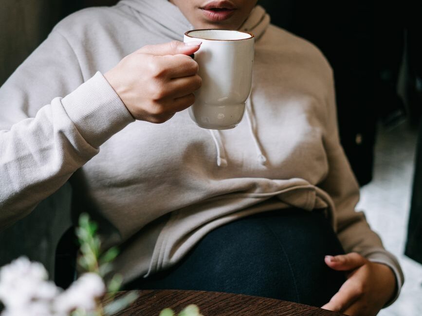 a pregnant woman touching her baby bump while drinking a mug of hot coffee
